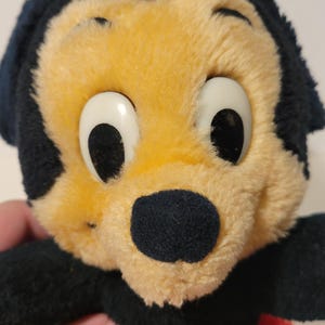 Puede incluir: Un juguete de peluche vintage con un personaje de dibujos animados con cabeza y orejas negras, cara amarilla y ojos blancos grandes. El juguete tiene una nariz negra y pantalones cortos rojos. El peluche es suave y parece bien conservado.