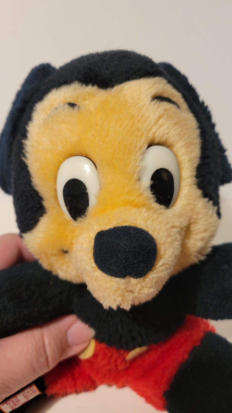 Puede incluir: Un juguete de peluche vintage con un personaje de dibujos animados con cabeza y orejas negras, cara amarilla y ojos blancos grandes. El juguete tiene una nariz negra y pantalones cortos rojos. El peluche es suave y parece bien conservado.