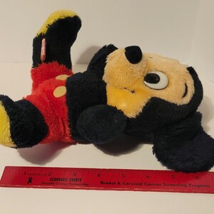 Puede incluir: Un peluche de Mickey Mouse, tumbado de lado. El juguete presenta un cuerpo negro, cara y pies amarillos, y pantalones cortos rojos con botones amarillos. El juguete mide aproximadamente 25 cm de largo.