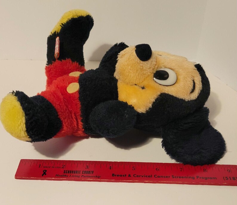Puede incluir: Un peluche de Mickey Mouse, tumbado de lado. El juguete presenta un cuerpo negro, cara y pies amarillos, y pantalones cortos rojos con botones amarillos. El juguete mide aproximadamente 25 cm de largo.