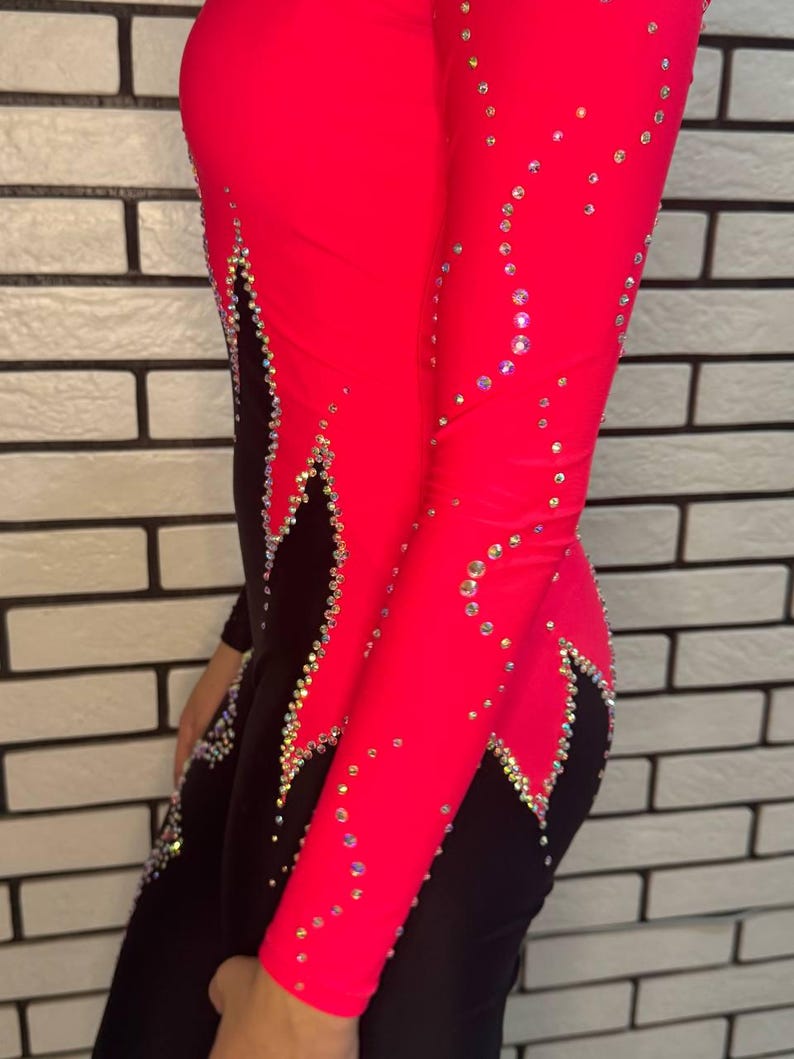Puede incluir: Un leotardo de manga larga con un dise&ntilde;o de bloques de color rosa intenso y negro. La prenda est&aacute; adornada con numerosos cristales transparentes y reflectantes dispuestos en patrones de remolinos a lo largo de las costuras y sobre la tela.