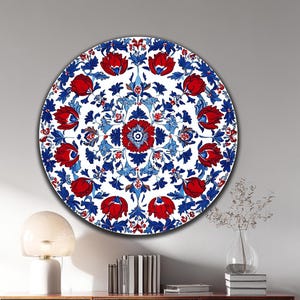 May include: A circular wall decoration with a floral pattern. It displays red flowers and blue foliage on a white base, enclosed by a blue border. The artwork is hung on a white wall above a wooden cabinet.
