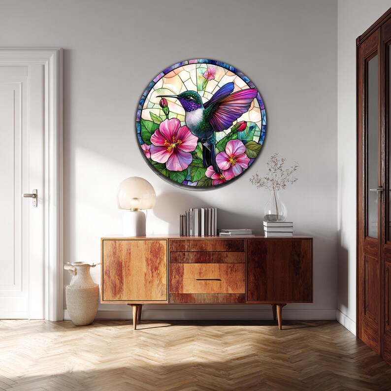 May include: A round stained glass art piece featuring a hummingbird with purple, green, and blue feathers, surrounded by pink hibiscus flowers and green leaves. The artwork is set against a light background and framed with a dark border. The piece is mounted on a white wall.