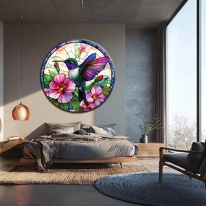 May include: A round wall art piece featuring a stained-glass design of a hummingbird and pink flowers. The hummingbird has green, purple, and white feathers. The flowers are a vibrant pink. The artwork is mounted on a wall above a bed.