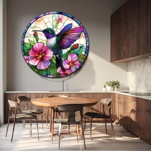 May include: Round stained glass art featuring a hummingbird with purple and green feathers, surrounded by pink hibiscus flowers and green leaves. The artwork is set against a backdrop of a light-colored, abstract design.