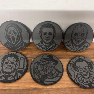 May include: Six grey slate coasters featuring engraved images of horror film characters, including Ghostface, Michael Myers, Jason Voorhees, Leatherface, Freddy Krueger, and Pennywise. The coasters are round and oval.
