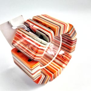 May include: A multi-colored striped bangle bracelet made of plastic. The bracelet is made up of several rectangular pieces that are joined together. The colors in the bracelet are orange, white, brown, and black.