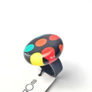 May include: A black ring with a large, oval-shaped top adorned with colorful polka dots in red, orange, yellow, teal, and blue. The ring band is a solid black color. The ring is on a white background.