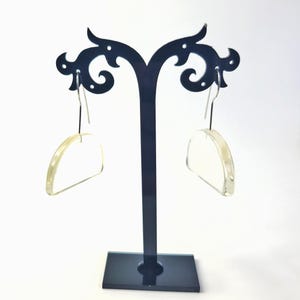 May include: A pair of clear, half-circle earrings with silver hooks are displayed on a black decorative earring stand. The earrings have a translucent appearance and a simple, modern design. The stand has a rectangular base and a curved top.