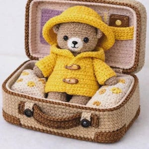 May include: A crocheted brown teddy bear wearing a yellow hooded raincoat with two wooden toggle buttons, sitting inside a tan crocheted suitcase. The suitcase interior features light-colored fabric cushions with small yellow floral patterns.