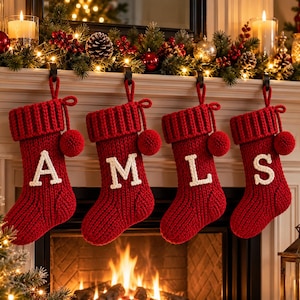 Può includere: Quattro calze di Natale in maglia rossa appese in fila. Ogni calza presenta un risvolto a coste, un pompon rosso abbinato e una grande lettera color crema cucita sulla parte anteriore. Le lettere visualizzate sulle calze sono A, M, L e S.
