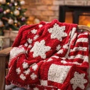 Peut inclure: Un plaid tricoté rouge et blanc orné d'un motif texturé de grands et petits flocons de neige blancs. La couverture est drapée sur une chaise en bois et présente une combinaison de sections en tricot torsadé et de panneaux carrés blancs.