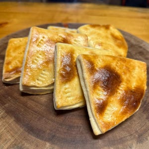 Puede incluir: Cinco pasteles de color marrón dorado, posiblemente empanadas, dispuestos en una tabla de cortar de madera. Los pasteles tienen forma triangular y una corteza escamosa y mantecosa.