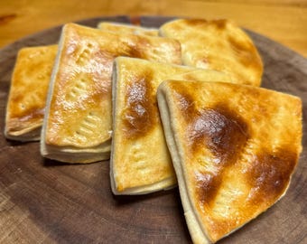 May include: Five golden brown pastries, possibly empanadas, arranged on a wooden cutting board. The pastries are triangular in shape and have a flaky, buttery crust.