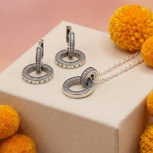 Può includere: Un paio di orecchini a cerchio in argento e una collana in argento abbinata, ciascuno con un ciondolo circolare inciso con la parola PANDORA e decorato con piccole pietre chiare. I gioielli sono esposti su una scatola beige con pon pon arancioni.