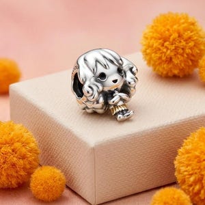 May include: A silver charm bead shaped like a stylized character with large eyes, wavy hair, and a small gold-toned skirt. The charm features a hollow center for a bracelet and is crafted with polished metallic surfaces and dark recessed detailing.