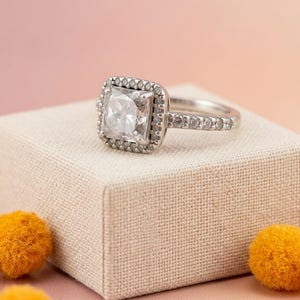 May include: A silver-toned ring featuring a square-cut centre stone surrounded by a halo of small, round, clear stones. The band is also set with a series of small, round, clear stones along the top half, creating a sparkling, multi-faceted appearance.