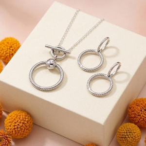 May include: A silver jewelry set consisting of a toggle necklace and two hoop earrings. The necklace features a chain with a circular pendant and a T-bar closure. The earrings are circular hoops with a twisted rope texture matching the pendant.