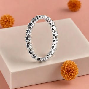 Op de afbeelding: Een zilveren ring met een herhalend sterrenpatroon. De ring wordt gepresenteerd op een lichtroze oppervlak, met oranje decoratieve bloemen op de achtergrond. Het ontwerp van de ring is gedetailleerd en ingewikkeld.