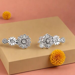 Puede incluir: Pendientes de plata con diseño de margaritas y detalles de cristal transparente. Cada pendiente presenta tres diseños de flores de margarita, con la flor más grande en el centro. Se exhiben sobre una superficie beige con acentos de flores amarillas.