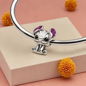 May include: A silver charm depicting the Disney character Stitch, with purple ear detailing, attached to a silver bracelet. The image includes yellow decorative balls in the background. The charm is a popular accessory.