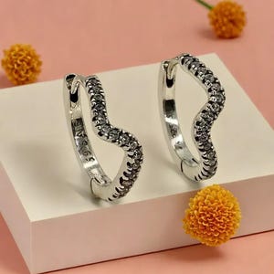 May include: A pair of silver-toned hoop earrings with a wavy design. The earrings are encrusted with small, clear stones along the outer curve. The earrings are displayed on a white surface with a pink background and orange floral accents.
