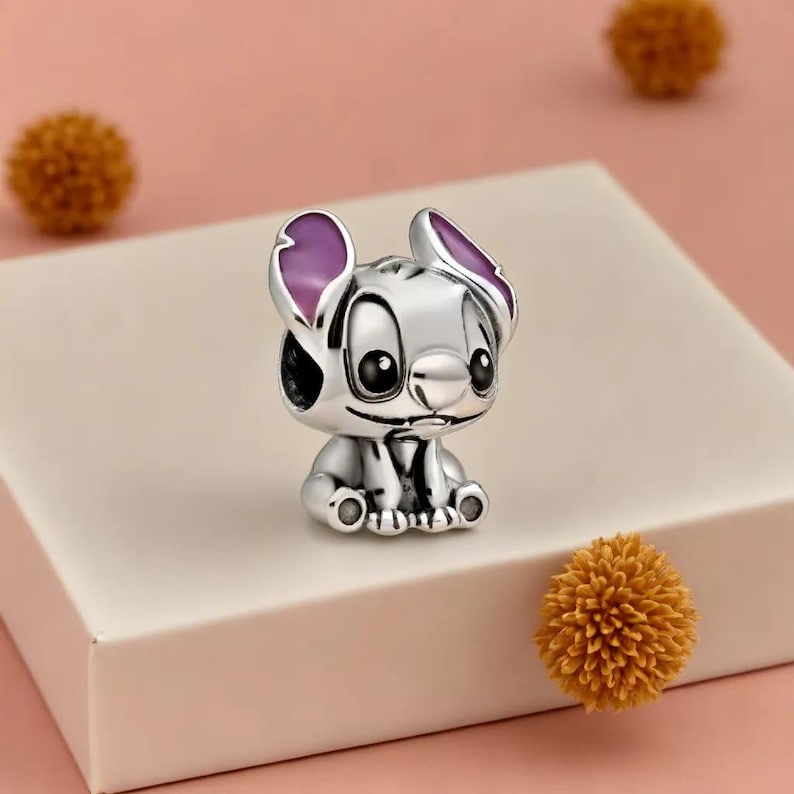 May include: A silver charm depicting a cartoon character with large black eyes and purple-lined ears. The charm is on a white surface with a pink background and two orange pom-poms.