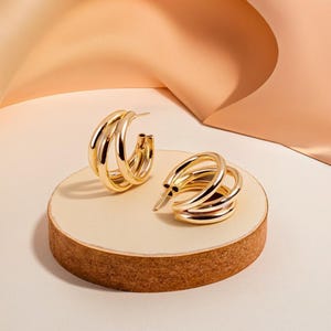 May include: A pair of gold hoop earrings with a triple layered design. The earrings are displayed on a small wooden platform.