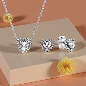 May include: A silver necklace and stud earrings set. Each piece has a heart-shaped, clear gemstone surrounded by smaller, clear stones. The jewellery is displayed on a beige surface with yellow flowers in the background.