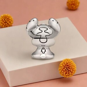 May include: A silver figurine of a cartoon character with large ears and a unique form. The figure has black detailing and is set against a light pink backdrop with yellow floral decorations.