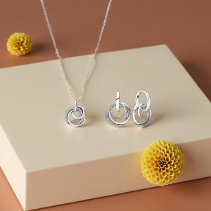 以下が含まれることがあります： 小さな輝く石で飾られた、連動する円が特徴のシルバーネックレスとイヤリングのセット。 ネックレスは繊細なチェーンで、イヤリングはフープスタイルです。 薄いベージュの表面に黄色い花が飾られています。