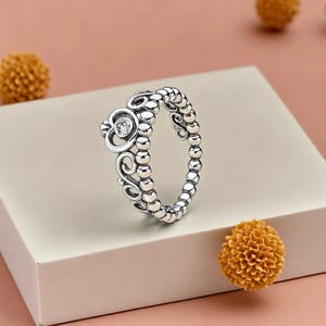 Pode incluir: Um anel de prata com um design de coroa. O anel apresenta uma banda de contas e um topo em forma de coroa com uma única pedra preciosa transparente. O anel é exibido em uma superfície branca.