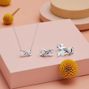 Peut inclure: Un ensemble de bijoux argentés avec un motif de symbole infini. L'ensemble comprend un collier et des boucles d'oreilles, chacun orné de petites pierres scintillantes. Les bijoux sont présentés sur une surface rose, avec des accents floraux jaunes.