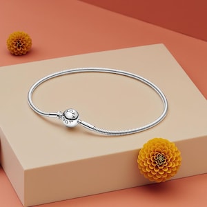 Op de afbeelding: Een zilveren armband met een slangenkettingontwerp. De armband heeft een ronde sluiting met de merknaam gegraveerd. Twee gele bloemen zijn geplaatst op een perzik- en beige achtergrond.