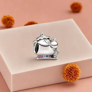 May include: A silver teapot charm with a smiling face, a small cup, and a small bottle. The charm has purple and teal accents on the base. The charm is set on a light pink surface. The charm is likely made of metal.