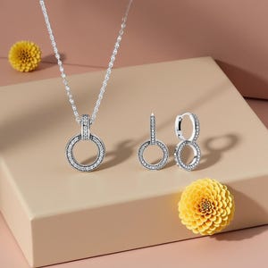 以下が含まれることがあります： 小さな輝く石がちりばめられた、円形のペンダントとフープが特徴のシルバーのネックレスとイヤリングのセット。ジュエリーは、2つの黄色い花と一緒にニュートラルな表面に展示されています。