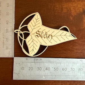 May include: A wooden leaf-shaped ornament with the name "Sean" etched into it. The ornament is approximately 7.6 cm long and features intricate leaf vein details. The design includes a circular frame around the leaf.