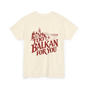 May include: A cream-coloured short-sleeve t-shirt with a speckled texture. The back features a dark red graphic illustration of a coastal village with a bell tower and a sailboat, accompanied by the text TOO BALKAN FOR YOU in a bold font.