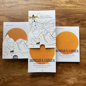 May include: Save the date card with a lighthouse illustration and mountains. The text reads "Meet Us Where Mountains Kiss Waves" and "Moriah & Lauren, October 4th, 2025, Isle Royale National Park, Moandlaurenmint.us".