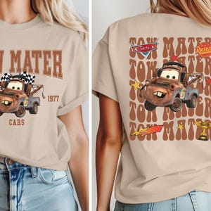 May include: Beige t-shirt featuring a graphic of Tow Mater from the Cars movie. The front design includes the text "TOW MATER EST. 1977 CARS". The back of the shirt has a larger image of Mater with the text "TOW MATER" repeated.