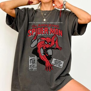 May include: A charcoal grey short-sleeve t-shirt featuring a vintage-style comic book graphic of Spider-Man in a crouching pose. The text reads: NEVER HAS ANYONE FOUGHT SUCH MERCILESS FOES AS... THE AMAZING SPIDER-MAN, FEATURING PETER PARKER ALIAS SPIDER-MAN.