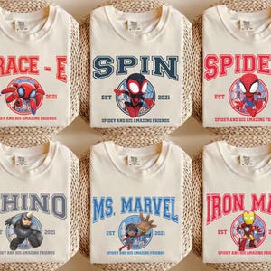 May include: Six cream-colored t-shirts featuring various Spider-Man characters and logos. Each shirt displays a different name like "Trace-E", "Spin", and "Spidey" in bold, collegiate-style lettering, along with character illustrations and the text "EST 2021".