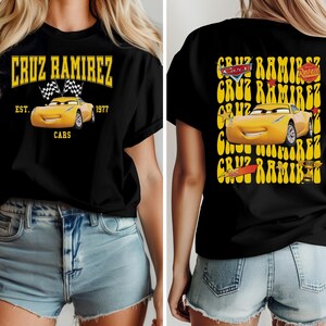 May include: Black t-shirt featuring a yellow graphic of Cruz Ramirez from the Cars movie. The front design includes the text "CRUZ RAMIREZ" and "CARS" with a yellow car and checkered flags. The back design repeats the name and car image.