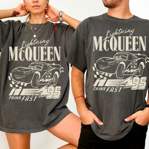 May include: Two charcoal grey t-shirts featuring a Lightning McQueen graphic. The design includes the text "Lightning McQueen", a cartoon car, the number "95", and the words "Think Fast". The shirts have short sleeves and a relaxed fit.