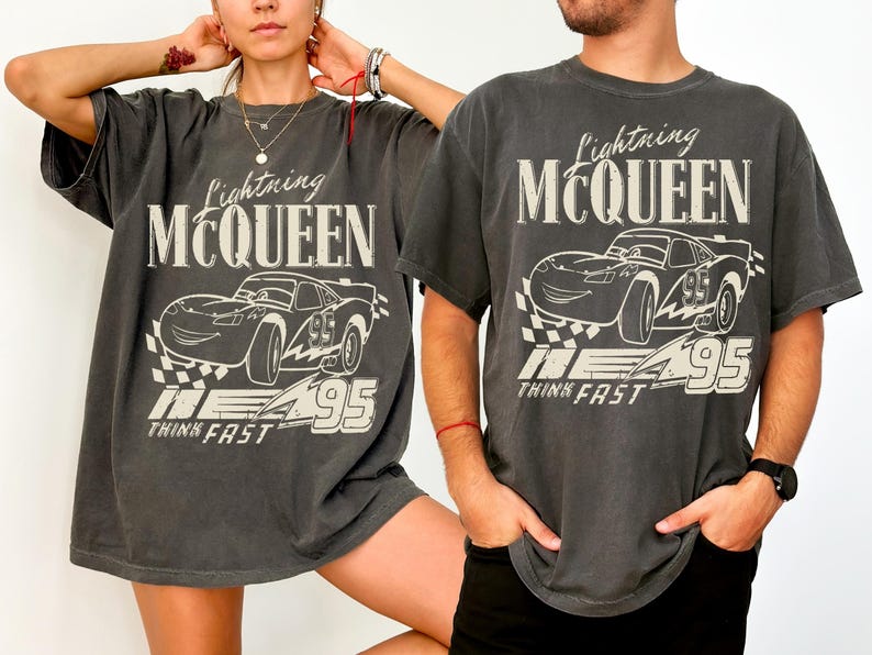 May include: Two charcoal grey t-shirts featuring a Lightning McQueen graphic. The design includes the text "Lightning McQueen", a cartoon car, the number "95", and the words "Think Fast". The shirts have short sleeves and a relaxed fit.