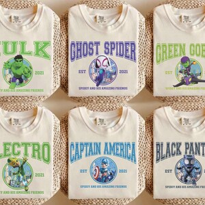 May include: Six cream-colored t-shirts featuring superhero graphics. Each shirt displays a different character's name in bold lettering, such as "Hulk" and "Ghost Spider", along with the text "EST 2021" and "Spidey and his Amazing Friends".