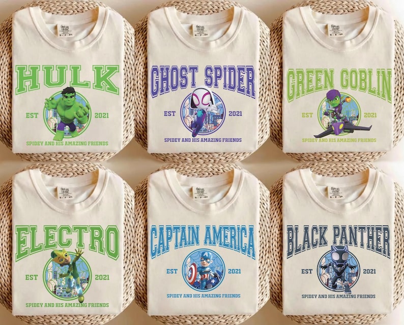May include: Six cream-colored t-shirts featuring superhero graphics. Each shirt displays a different character's name in bold lettering, such as "Hulk" and "Ghost Spider", along with the text "EST 2021" and "Spidey and his Amazing Friends".