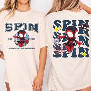 May include: Cream-colored t-shirt featuring a cartoon Spider-Man design. The front of the shirt displays the word "SPIN" above a graphic of Spider-Man, with "EST 2021" and "SPIDEY AND HIS AMAZING FRIENDS" below. The back has a larger "SPIN" design with Spider-Man and stars.