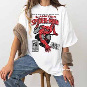 May include: A white t-shirt featuring a vintage comic book graphic of Spider-Man in a red and black suit. The text reads: NEVER HAS ANYONE FOUGHT SUCH MERCILESS FOES AS... THE AMAZING SPIDER-MAN, FEATURING PETER PARKER ALIAS SPIDER-MAN, AND ANOTHER SPECTACULAR SMASH-HIT FROM THE HOUSE OF IDEAS!