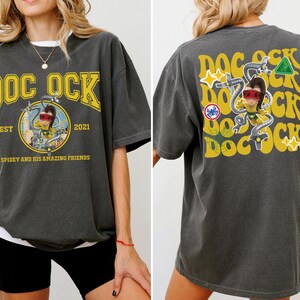 May include: Dark gray t-shirt with a yellow "DOC OCK" graphic on the front and back. The front graphic includes a circular image of a cartoon character and the text "EST 2021" and "SPIDEY AND HIS AMAZING FRIENDS". The back has a repeating "DOC OCK" design.